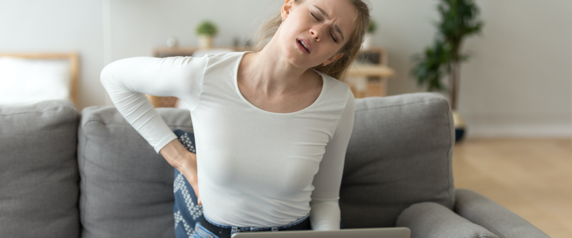 mulher sentada no sofá de frente a um computador com as mãos nas costas com expressão de dor